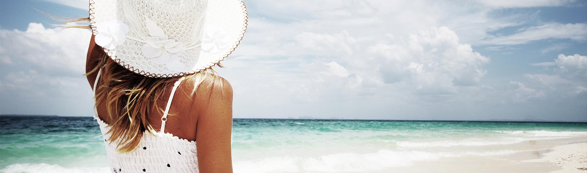 A woman holding onto her white hat during a day at the beach.